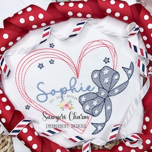 May include: Embroidered design featuring a red, white, and blue bow with stars, a heart outline with red stitching, and the name "Sophie" with the text "Est. 2021 Dothan" below. The design is on a white fabric background.