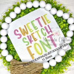 May include: A white square with the words "SWEETIE SKETCH FONT" in colorful, stitched lettering. The design is surrounded by a green wreath with white beads and a brown tassel.