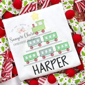 May include: A green toy train shaped like a Christmas tree with a yellow star on top. The train has red wheels and is decorated with white squares. The name "HARPER" is embroidered below the train.