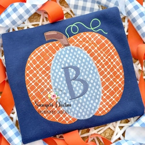 May include: Navy blue shirt with an embroidered pumpkin design. The pumpkin is orange with a white grid pattern, a brown stem, and a light blue circle with the letter "B". The shirt has the text "Sawyers Charm Embroidery Designs".