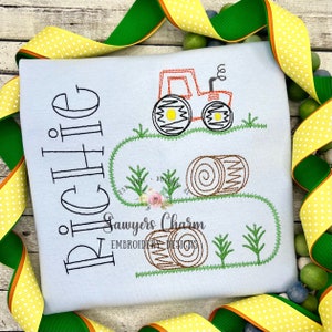 May include: A light blue t-shirt with a black embroidered design of a tractor pulling hay bales. The text "Sawyers Charm Embroidery Designs" is also embroidered on the shirt. The name "RICHIE" is embroidered in a large, cursive font on the left side of the shirt.