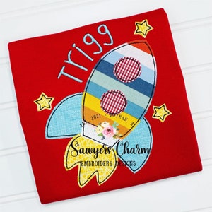 Puede incluir: Camiseta roja con una aplicación de cohete azul, amarillo y rojo. El cohete tiene un patrón a cuadros y el texto "Sawyers Charm Embroidery Designs" está debajo del cohete. El nombre "Trigg" está bordado en azul en la camiseta.