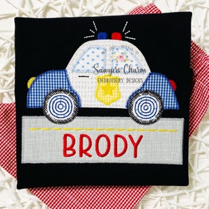May include: A black t-shirt with an embroidered design of a blue and white police car with a yellow star on the side. The car is on a gray road with the name "BRODY" in red letters below it.