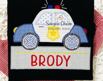 May include: A black t-shirt with an embroidered design of a blue and white police car with a yellow star on the side. The car is on a gray road with the name "BRODY" in red letters below it.