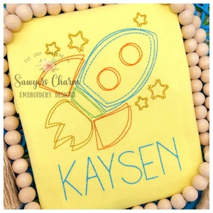 May include: Yellow embroidered design of a rocket ship with stars and the name Kaysen. The design is on a yellow fabric.