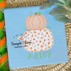 Puede incluir: Una camiseta azul claro con un diseño bordado de dos calabazas apiladas. La calabaza de arriba es de rayas naranja y blanca, y la calabaza de abajo es blanca con lunares naranja. El texto "Sawyers Charm Embroidery Designs" y "EST. 2021 - DOTRAN, AL" están bordados en la camiseta. El nombre "Adley" está bordado en verde debajo de las calabazas.