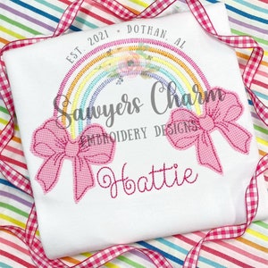 May include: White t-shirt with a rainbow design, two pink gingham bows, and the name "Hattie" embroidered in pink. The text "Sawyers Charm Embroidery Designs" and "Est. 2021 Dothan, AL" are also visible.