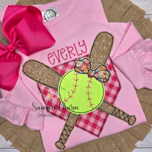 Op de afbeelding: Roze shirt met lange mouwen met een softbalontwerp. Het ontwerp omvat een neongele softbal, gekruiste houten knuppels en een bloemenstrik. De naam "Everly" is boven het ontwerp geborduurd. De shirt heeft een geplooid mouwtje en een grote roze strik.