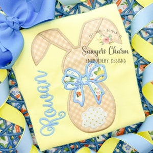May include: A yellow t-shirt with a brown bunny applique with a blue bow. The bunny has a gingham pattern and a white polka dot tail. The shirt also has the name "Rowan" embroidered in blue thread. The shirt has the text "Sawyers Charm Embroidery Designs" printed on it.