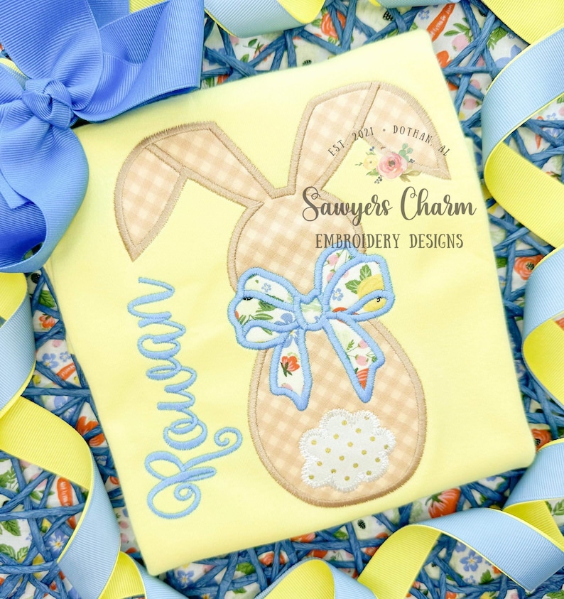 May include: A yellow t-shirt with a brown bunny applique with a blue bow. The bunny has a gingham pattern and a white polka dot tail. The shirt also has the name "Rowan" embroidered in blue thread. The shirt has the text "Sawyers Charm Embroidery Designs" printed on it.