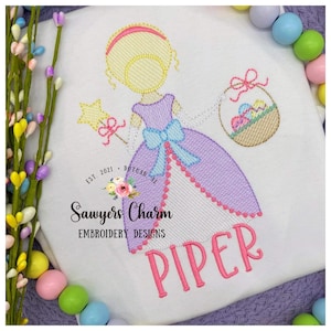 May include: Embroidered design of a girl in a purple dress holding a basket of Easter eggs. The girl has blonde hair and is wearing a blue bow. The design includes the text "Piper" in pink lettering.