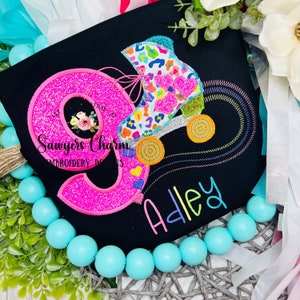 May include: A black t-shirt with pink glitter number nine, a colorful roller skate, and the name "Adley" embroidered in rainbow colors. The design is on a black t-shirt.