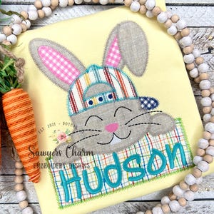 以下が含まれることがあります： 黄色のTシャツに、ストライプの帽子をかぶったウサギが箱から顔を出している刺繍のデザインが施されています。箱には「Hudson」という名前が刺繍されています。Tシャツには「Sawyers Charm Embroidery Designs」という刺繍も施されています。