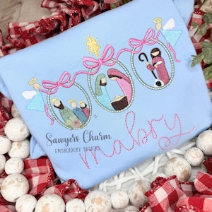 May include: A light blue long-sleeve shirt featuring embroidered Christmas ornaments with angels, a star, and nativity figures. The name "Malory" is stitched in pink script. The shirt also has the text "Sawyers Charm Embroidery Designs."