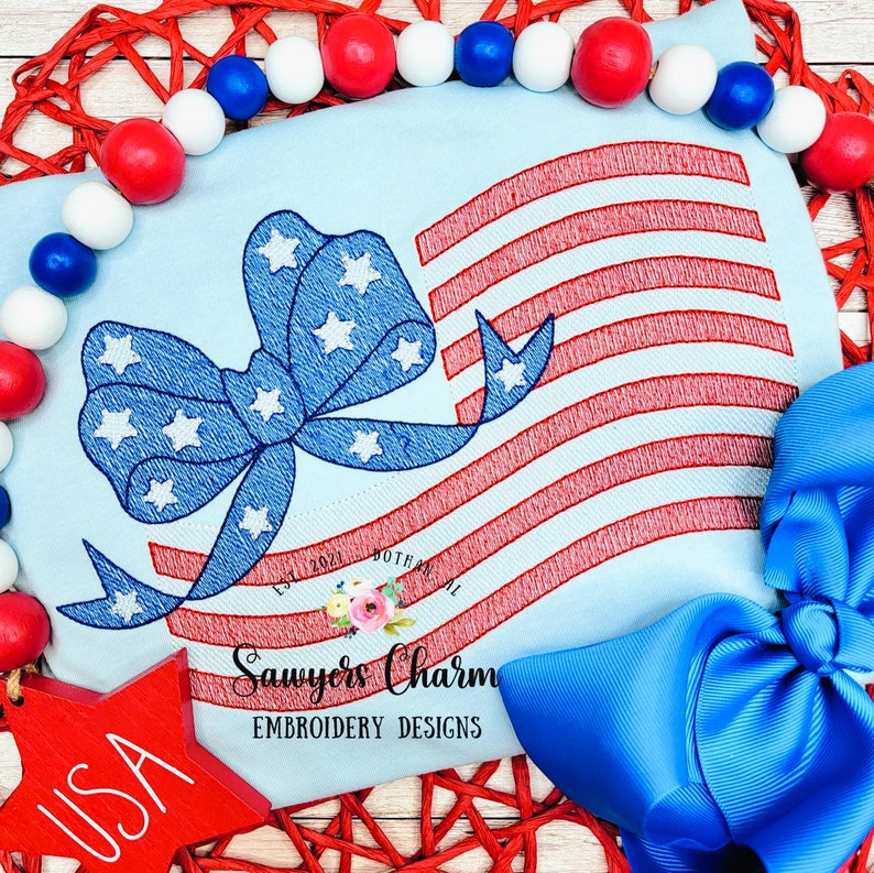 May include: Embroidered design of an American flag with a blue bow with white stars. The design is on a light blue fabric. The text "Sawyer's Charm Embroidery Designs" is below the design.
