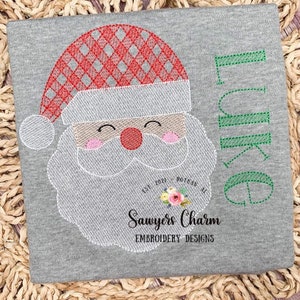 May include: Embroidered design of a Santa Claus face with a red and white checkered hat, a white beard, and a red nose. The design includes the text "Sawyers Charm Embroidery Designs" and "Est. 2021 Bozeman, MT".