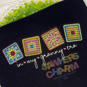 May include: Black fabric with embroidered squares in vibrant colours. The text reads "in my granny era" and "Sawyers Charm". The squares feature floral and geometric designs in pink, yellow, blue, and red.