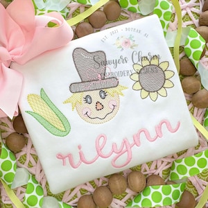 以下が含まれることがあります： ピンクの刺繍で「rilynn」という名前が書かれた白いロンパース。かわいい刺繍の案山子とひまわり、トウモロコシが描かれています。ロンパースには「Sawyers Charm Embroidery Designs」という刺繍も入っています。
