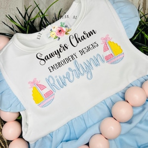 May include: A white baby onesie with blue trim and embroidered text that reads "Sawyers Charm Embroidery Designs Riverlynn". The onesie features two embroidered images of sailboats with pink, yellow, and blue accents.