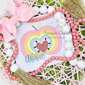 May include: A pink bow and beaded garland surround a white square with an embroidered apple design. The apple features a rainbow pattern, a red heart, and the name "Abbie" in teal script. The design is from "Sawyers Charm Embroidery Designs."