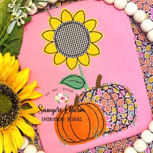 以下が含まれることがあります： 刺繍されたひまわりと2つのカボチャ、そして「Sawyers Charm Embroidery Designs Est. 2021 Dothan, AL」というテキストが書かれたピンクのTシャツです。