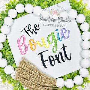 May include: A white tag with the text "The Bougie Font" in colorful embroidery. The tag is surrounded by a green wreath, white beads, and a bundle of twine. The text "Sawyers Charm Embroidery Designs" is also visible.