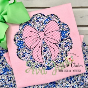May include: Pink and white floral fabric with a large pink bow applique. The bow is outlined in black and has a white gingham pattern. The fabric has a scalloped edge. The text "Ava" and "Sawyers Charm Embroidery Designs" is embroidered on the fabric.