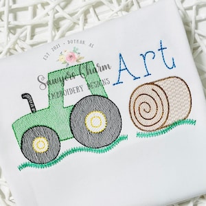 Puede incluir: Tractor verde con una paca de heno y el nombre Art bordado en una camisa blanca. El tractor tiene una rueda amarilla y una paca de heno marrón.