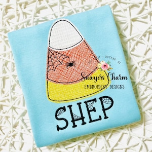 May include: A light blue t-shirt with an embroidered candy corn design. The candy corn has a spider web and a spider on it. The text "SHEP" is embroidered below the candy corn.