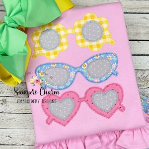 May include: Three pairs of sunglasses with different shapes and colors. The sunglasses are appliqued on a pink fabric with a ruffle trim. The sunglasses are yellow, blue, and pink. The lenses are gray with white polka dots.