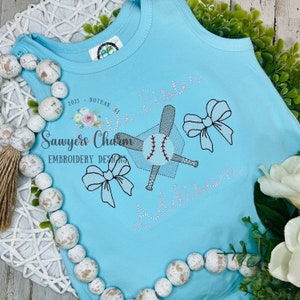 May include: A light blue tank top with a baseball and bat design, two bows, and the text "Sawyers Charm Embroidery Designs" and "Big Sister Addison".