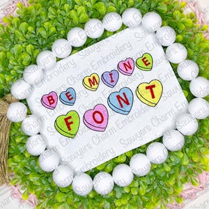 Puede incluir: Diseño bordado de la frase "Be Mine" en una fuente de corazón de caramelo colorido. Las letras están cosidas en rosa, azul, verde, amarillo y rojo sobre un fondo blanco.