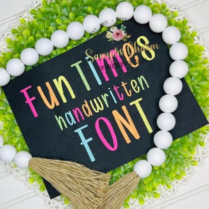May include: A black sign with the words "Fun Times handwritten FONT" in colorful, handwritten-style lettering. The sign is surrounded by a white beaded garland and green foliage.