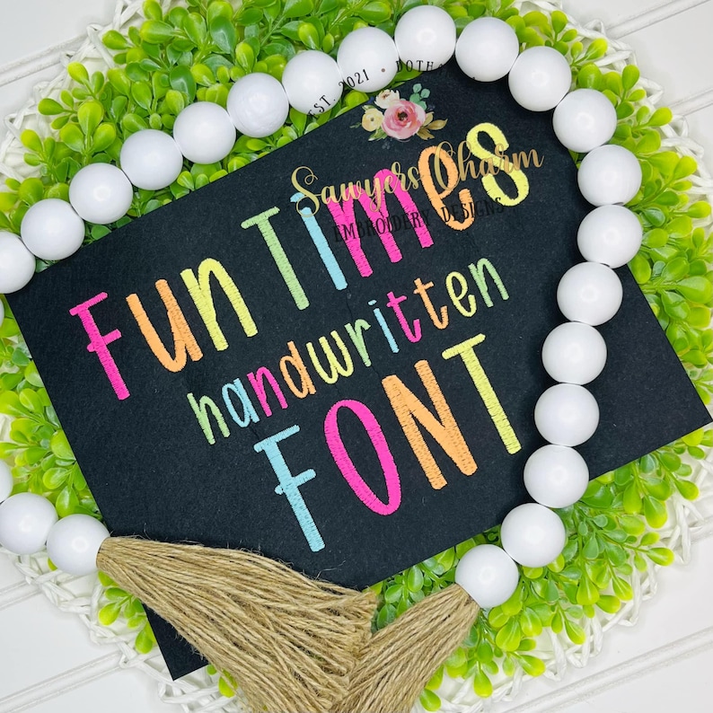 May include: A black sign with the words "Fun Times handwritten FONT" in colorful, handwritten-style lettering. The sign is surrounded by a white beaded garland and green foliage.