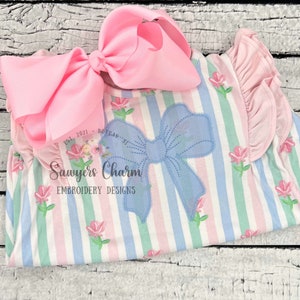 Könnte beinhalten: Ein blau-weiß gestreiftes Kleid mit einem hellblauen Schleifen-Applik. Das Kleid hat rosa Rüschen an den Ärmeln und eine rosa Schleife an der Schulter. Das Kleid hat einen Blumendruck mit rosa Blumen. Der Text "Sawyers Charm Embroidery Designs" ist auf das Kleid gedruckt.