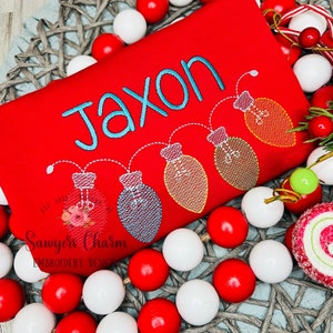 Puede incluir: Camiseta roja de Navidad con el nombre Jaxon bordado en azul turquesa. La camiseta presenta una cadena de cinco bombillas de luz azul turquesa, roja y amarilla.