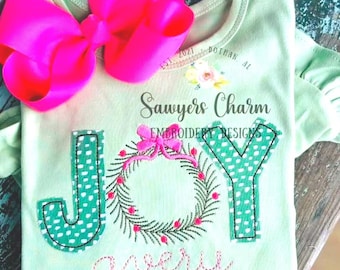 Op de afbeelding: Een lichtgroene babybody met het woord "JOY" in groene letters met witte stippen. Tussen de letters is een krans met een roze strik geborduurd. De naam "avery" is eronder geborduurd. Een grote roze strik is bevestigd.