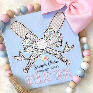 May include: A light blue t-shirt with a floral bow and two baseball bats. The bats are brown and white gingham. The bow is pink, white, and blue. The text "Sawyer's Charm Embroidery Designs" and "Rylie Ann" is embroidered on the shirt.