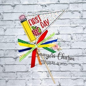 Può includere: Un gagliardetto decorativo con la scritta "FIRST DAY" e un'illustrazione di una mela. Una matita gialla è sul lato sinistro. Il gagliardetto è decorato con nastri colorati e un bastoncino di legno. Il design è di Sawyer's Charm Embroidery Designs.