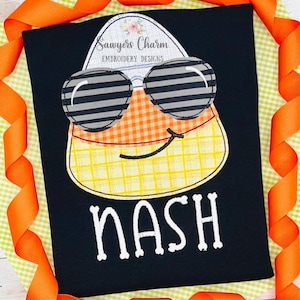 May include: A black t-shirt with an embroidered image of a candy corn wearing sunglasses. The candy corn is yellow, orange, and white with a checkered pattern. The text "Sawyers Charm Embroidery Designs" and the name "NASH" are also embroidered on the shirt.