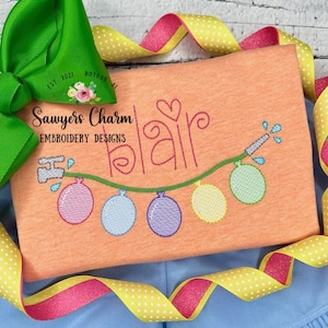 May include: A light orange t-shirt with the name "Blair" embroidered in pink thread with a heart above the name. A green line with a water sprinkler at the beginning and five colorful balloons hanging from the line.