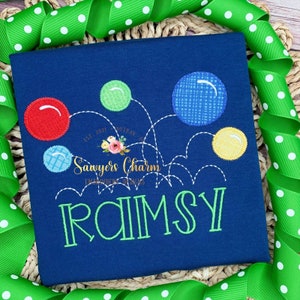 以下が含まれることがあります： 緑、赤、青、黄色の風船のデザインが施された紺色のTシャツ。シャツには「Sawyers Charm Embroidery Designs」と「EST. 2021 DOTHAN, AL」の文字が刺繍されている。シャツの下部には緑色の文字で「RAMSY」と刺繍されている。