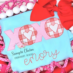 May include: A light blue t-shirt with a red and white checkered XO design and a white heart in each circle. The shirt also has the name "emory" embroidered in red cursive script below the XO design. The shirt is surrounded by pink and red ribbon.