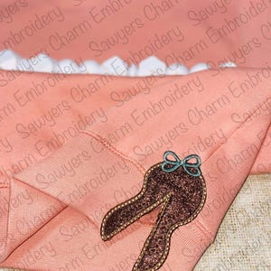 May include: A pink sweatshirt with a brown glitter bunny ear applique. The sweatshirt has the text "Sawyers Charm Embroidery" repeated on the fabric.