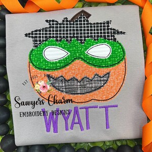 Puede incluir: Una camiseta gris con un diseño bordado de una calabaza con una máscara verde. La calabaza tiene una boca cosida negra y un sombrero cosido negro. El texto "Sawyers Charm Embroidery Designs" y el nombre "WYATT" están bordados en morado debajo de la calabaza.