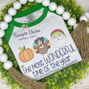 May include: White and green raglan style t-shirt with embroidered images of a pumpkin, turkey, and Santa Claus. The text "The Most Wonderful Time of the Year" is embroidered below the images. The shirt is size 3T.