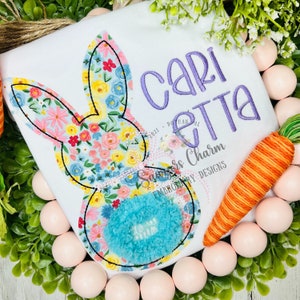 May include: A white onesie with a floral bunny applique and the name "Caritta" embroidered in purple. The onesie is surrounded by pink Easter eggs and a carrot.
