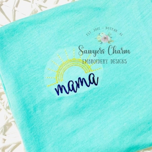 May include: A turquoise t-shirt with an embroidered design of a yellow sun with a rainbow arc and the word "mama" in blue cursive lettering. The text "Sawyers Charm Embroidery Designs" is also embroidered on the shirt.