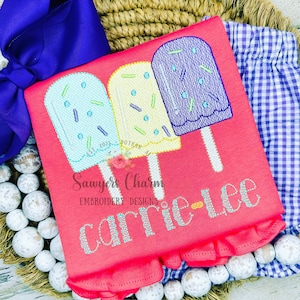 Puede incluir: Una camiseta rosa con un dobladillo con volantes presenta un diseño bordado de tres paletas de hielo con chispas. Las paletas de hielo son amarillas, azules y verdes. El texto "Sawyer's Charm Embroidery Designs" y "carrie-lee" también está bordado en la camiseta.