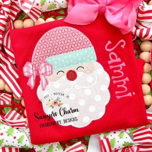 May include: Red Christmas shirt with a white Santa face applique with pink and blue accents. The shirt is personalized with the name "Sammy!" in pink thread. The shirt also has the text "Sawyers Charm Embroidery Designs Est. 2021 Dothan, AL" in black thread.